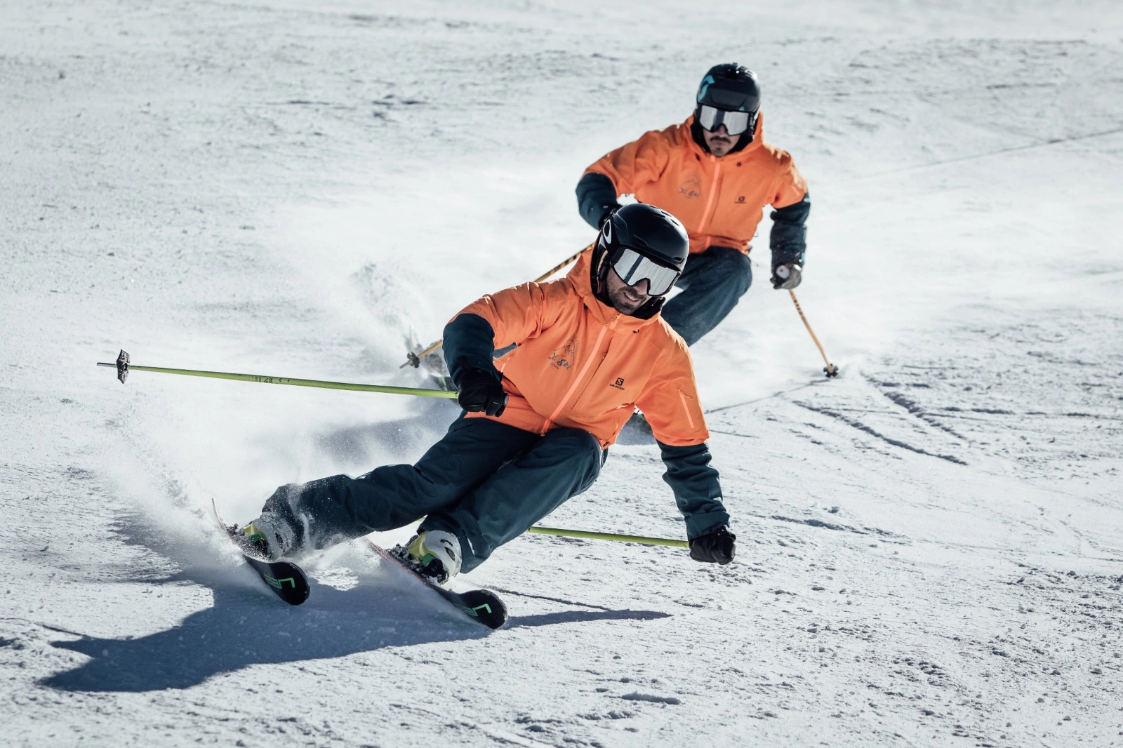 Dos hombres vestidos con equipación de esquí, azul y naranja, bajando con esquís por una montaña a alta velocidad